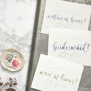 May include: Three cards with calligraphy text on white cardstock. The text on the cards reads "Will you be my matron of honor?", "Will you be my bridesmaid?", and "Will you be my maid of honor?"