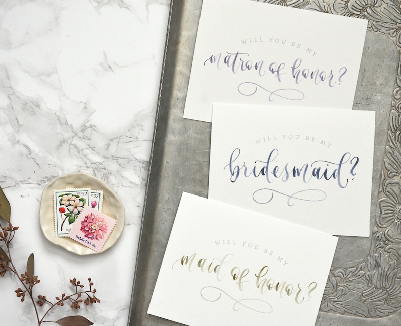 May include: Three cards with calligraphy text on white cardstock. The text on the cards reads "Will you be my matron of honor?", "Will you be my bridesmaid?", and "Will you be my maid of honor?"