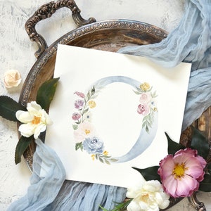 May include: A watercolor painting of a floral letter O. The letter is painted in a light blue color and surrounded by a wreath of pink, purple, and yellow flowers. The background is white.