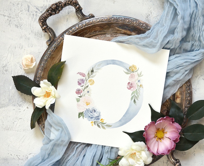 May include: A watercolor painting of a floral letter O. The letter is painted in a light blue color and surrounded by a wreath of pink, purple, and yellow flowers. The background is white.