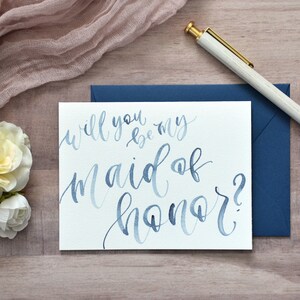 Puede incluir: Una tarjeta blanca con caligraf&iacute;a azul en acuarela que dice "&iquest;Quieres ser mi dama de honor?" La tarjeta est&aacute; sobre una superficie de madera con un sobre azul y un bol&iacute;grafo blanco.
