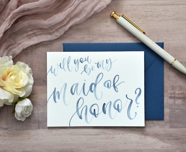 Puede incluir: Una tarjeta blanca con caligraf&iacute;a azul en acuarela que dice "&iquest;Quieres ser mi dama de honor?" La tarjeta est&aacute; sobre una superficie de madera con un sobre azul y un bol&iacute;grafo blanco.