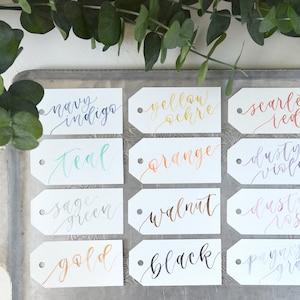 Puede incluir: Doce etiquetas blancas con nombres de colores escritos a mano en varios tonos de tinta. Los colores incluyen azul &iacute;ndigo, verde azulado, verde salvia, dorado, negro, ocre amarillo, naranja, rojo escarlata, violeta polvoriento, rosa polvoriento y gris Payne.