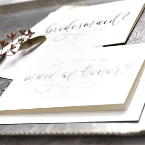 May include: Two white cards with gray calligraphy. The top card says "bridesmaid?" and the bottom card says "Will you be my maid of honor?"