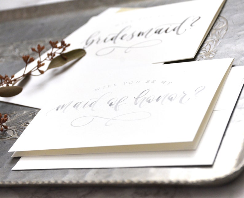 May include: Two white cards with gray calligraphy. The top card says "bridesmaid?" and the bottom card says "Will you be my maid of honor?"