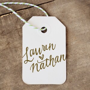 May include: White gift tag with a green and white twine string attached. The tag has the names "Lauren" and "Nathan" written in cursive with a small heart symbol between the names.