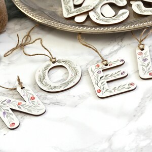 May include: Wooden Christmas ornaments spelling out the word "NOEL" in white letters with green leaves and red and purple berries painted on them.