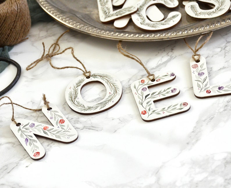 May include: Wooden Christmas ornaments spelling out the word "NOEL" in white letters with green leaves and red and purple berries painted on them.