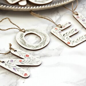 May include: Wooden Christmas ornaments with a floral design. The ornaments spell out the word "NOEL" with a wreath in place of the "O".