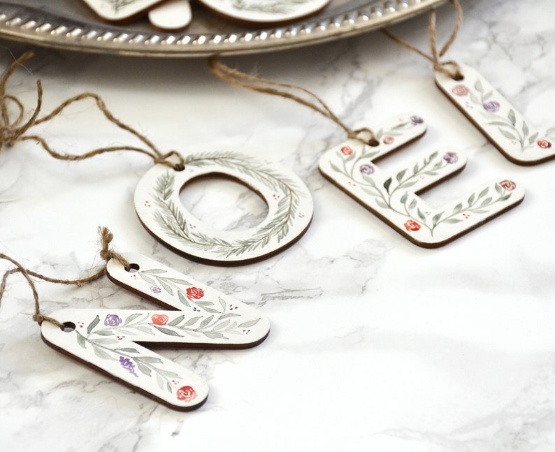 May include: Wooden Christmas ornaments with a floral design. The ornaments spell out the word "NOEL" with a wreath in place of the "O".