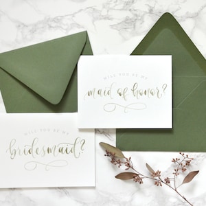 May include: Two white cards with gold calligraphy on a marble background. The top card says "Will you be my maid of honor?" and the bottom card says "Will you be my bridesmaid?"  The cards are surrounded by green envelopes and a sprig of dried flowers.