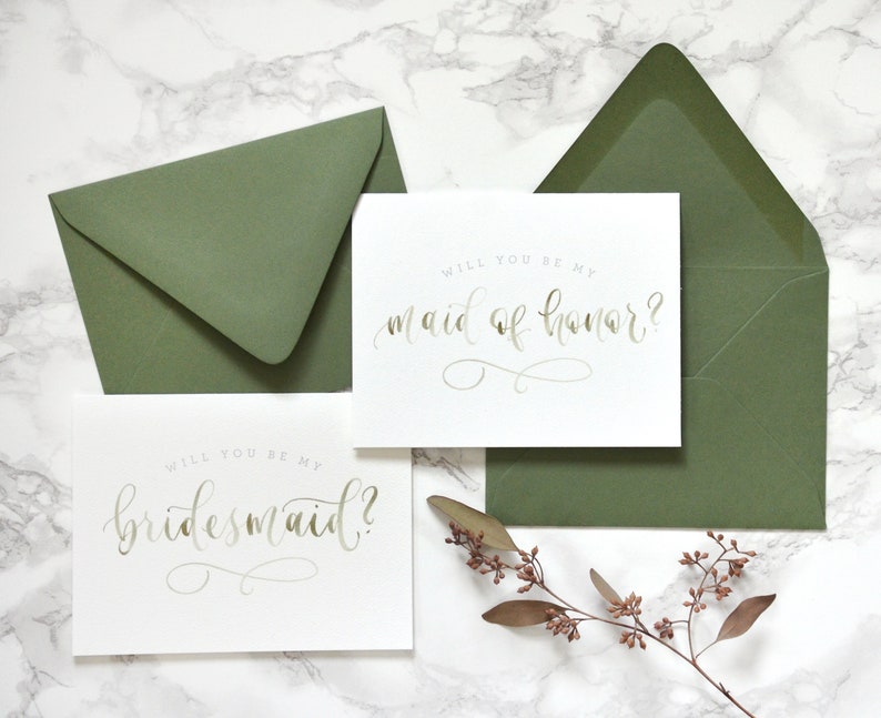 May include: Two white cards with gold calligraphy on a marble background. The top card says "Will you be my maid of honor?" and the bottom card says "Will you be my bridesmaid?"  The cards are surrounded by green envelopes and a sprig of dried flowers.