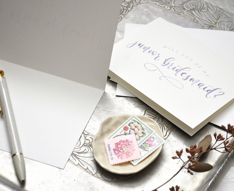 May include: A white card with the text "Will you be my junior bridesmaid?" written in purple calligraphy. The card is on a silver tray with a small plate of vintage stamps.