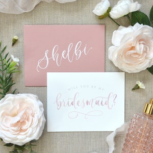May include: A pink card with the text "Shelbi" in white calligraphy. A white card with the text "Will you be my bridesmaid?" in pink calligraphy.