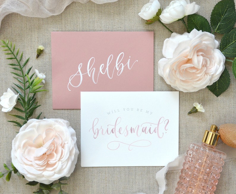 May include: A pink card with the text "Shelbi" in white calligraphy. A white card with the text "Will you be my bridesmaid?" in pink calligraphy.