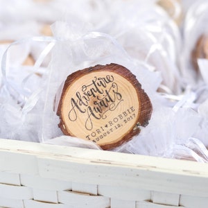 Puede incluir: Un peque&ntilde;o c&iacute;rculo de madera con el texto "Adventure Awaits" en escritura cursiva. El c&iacute;rculo est&aacute; decorado con un coraz&oacute;n y el texto "Tori & Robbie August 12, 2017". El c&iacute;rculo est&aacute; en una bolsa de organza blanca.