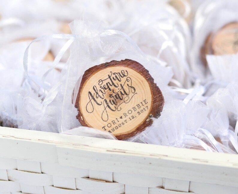 Puede incluir: Un peque&ntilde;o c&iacute;rculo de madera con el texto "Adventure Awaits" en escritura cursiva. El c&iacute;rculo est&aacute; decorado con un coraz&oacute;n y el texto "Tori & Robbie August 12, 2017". El c&iacute;rculo est&aacute; en una bolsa de organza blanca.