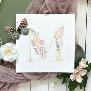 May include: Watercolor painting of the letter M decorated with pink and white flowers and green leaves. A butterfly is in the upper left corner of the image.