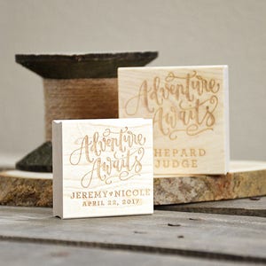 Puede incluir: Dos sellos de madera con el texto "Adventure Awaits" en una fuente cursiva. El sello superior tiene el texto "Shepard Judge" debajo del texto cursivo. El sello inferior tiene el texto "Jeremy & Nicole April 22, 2017" debajo del texto cursivo.