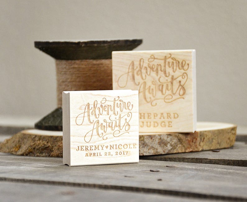 Puede incluir: Dos sellos de madera con el texto "Adventure Awaits" en una fuente cursiva. El sello superior tiene el texto "Shepard Judge" debajo del texto cursivo. El sello inferior tiene el texto "Jeremy & Nicole April 22, 2017" debajo del texto cursivo.