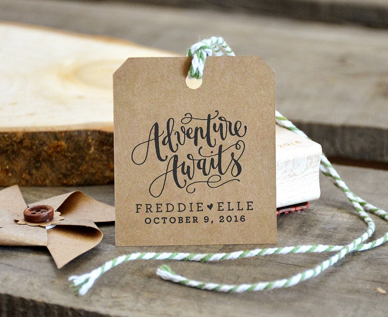 Puede incluir: Una etiqueta de papel marr&oacute;n con el texto "Adventure Awaits" en escritura cursiva negra. Debajo del texto se encuentra "Freddie &hearts; Elle October 9, 2016" en negro.