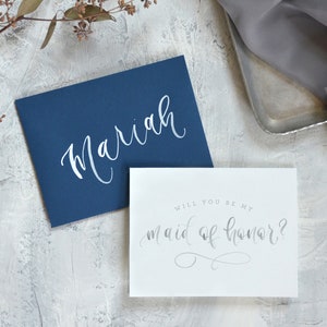 May include: A white card with the text "Will you be my maid of honor?" in silver script. The card is on a gray background with a blue envelope with the name "Mariah" written in white script.