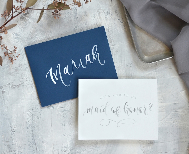 May include: A white card with the text "Will you be my maid of honor?" in silver script. The card is on a gray background with a blue envelope with the name "Mariah" written in white script.