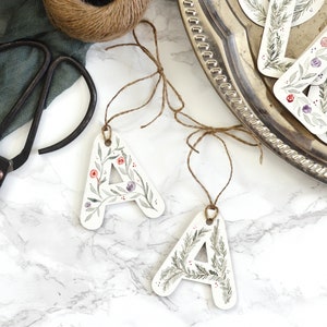 May include: Three white paper gift tags with floral and greenery designs. The tags are shaped like the letter A and have twine attached. The tags are on a white marble surface.