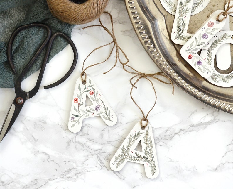 May include: Three white paper gift tags with floral and greenery designs. The tags are shaped like the letter A and have twine attached. The tags are on a white marble surface.