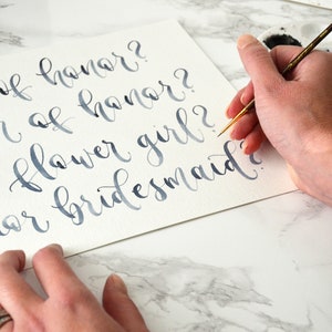 May include: A hand writes in blue ink on a white card with a calligraphy pen. The card says "Maid of honor?", "Matron of honor?", "Flower girl?", and "Junior bridesmaid?"