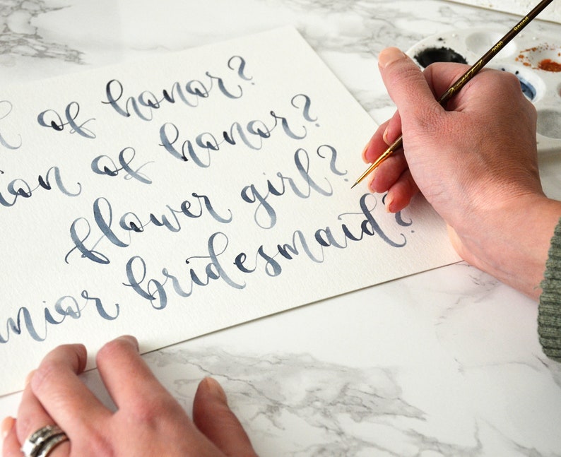 May include: A hand writes in blue ink on a white card with a calligraphy pen. The card says "Maid of honor?", "Matron of honor?", "Flower girl?", and "Junior bridesmaid?"