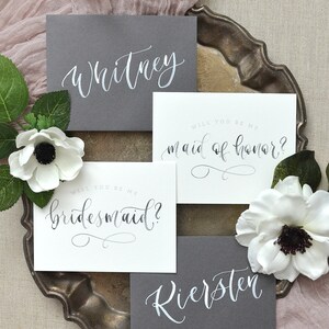 May include: Two gray cards with white calligraphy that reads "Whitney" and "Riersten". Two white cards with black calligraphy that reads "Will you be my bridesmaid?" and "Will you be my maid of honor?"