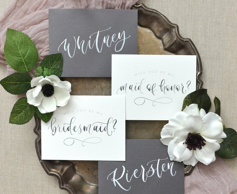 May include: Two gray cards with white calligraphy that reads "Whitney" and "Riersten". Two white cards with black calligraphy that reads "Will you be my bridesmaid?" and "Will you be my maid of honor?"