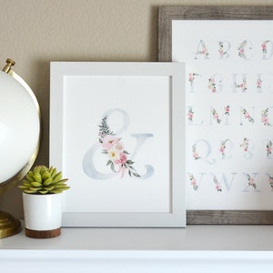 May include: Two framed prints with watercolor floral designs. The left print features the ampersand symbol with pink and white flowers. The right print features the alphabet with floral designs.
