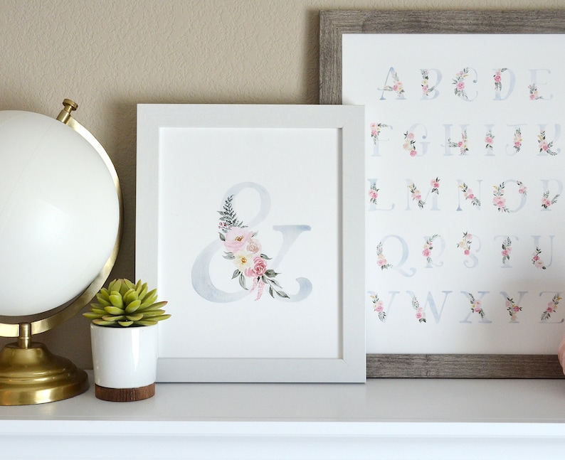 May include: Two framed prints with watercolor floral designs. The left print features the ampersand symbol with pink and white flowers. The right print features the alphabet with floral designs.