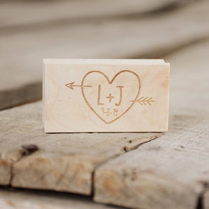 May include: A wooden block with a heart design engraved with the initials "L+J" and the date "9-8-14". An arrow points to the left of the heart and another arrow points to the right of the heart.