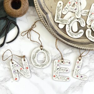 May include: Wooden ornaments spelling out the word "NOEL" with a floral design in shades of green, purple, and red. The ornaments are hanging from twine and are displayed on a white surface.