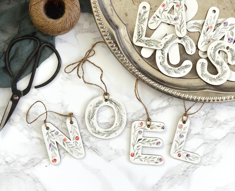 May include: Wooden ornaments spelling out the word "NOEL" with a floral design in shades of green, purple, and red. The ornaments are hanging from twine and are displayed on a white surface.