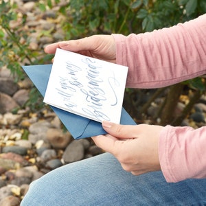 Puede incluir: Una tarjeta blanca con caligraf&iacute;a azul que dice "&iquest;Quieres ser mi dama de honor?" La tarjeta la est&aacute; sosteniendo una persona que lleva una camisa rosa y unos vaqueros azules.