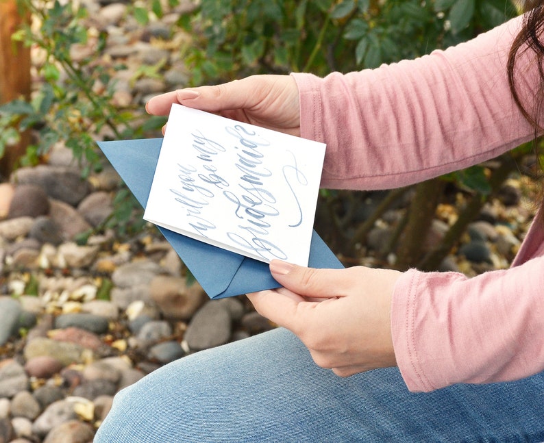 Puede incluir: Una tarjeta blanca con caligraf&iacute;a azul que dice "&iquest;Quieres ser mi dama de honor?" La tarjeta la est&aacute; sosteniendo una persona que lleva una camisa rosa y unos vaqueros azules.