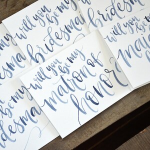 Puede incluir: Cinco tarjetas blancas con caligraf&iacute;a azul en acuarela. Las tarjetas dicen "&iquest;Quieres ser mi dama de honor?", "&iquest;Quieres ser mi madrina de honor?" y "&iquest;Quieres ser mi dama de honor?"