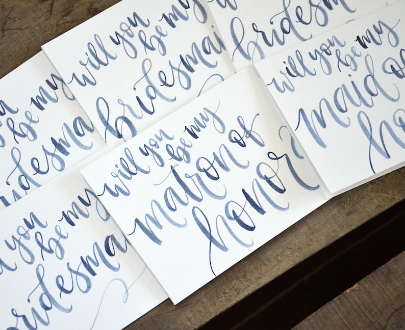Puede incluir: Cinco tarjetas blancas con caligraf&iacute;a azul en acuarela. Las tarjetas dicen "&iquest;Quieres ser mi dama de honor?", "&iquest;Quieres ser mi madrina de honor?" y "&iquest;Quieres ser mi dama de honor?"