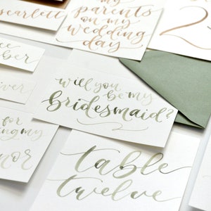 Puede incluir: Un conjunto de tarjetas de caligraf&iacute;a con tinta verde y dorada. Las tarjetas presentan frases como "&iquest;Quieres ser mi dama de honor?" y "Mesa doce".