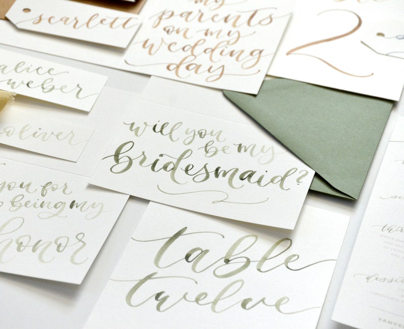 Puede incluir: Un conjunto de tarjetas de caligraf&iacute;a con tinta verde y dorada. Las tarjetas presentan frases como "&iquest;Quieres ser mi dama de honor?" y "Mesa doce".