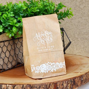 Puede incluir: Una bolsa de papel marr&oacute;n con texto blanco que dice "Adventure Awaits" y "Olivia June Due June 2017". La bolsa tiene un dise&ntilde;o floral blanco en la parte inferior.