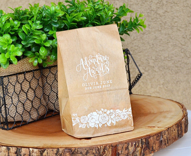 Puede incluir: Una bolsa de papel marr&oacute;n con texto blanco que dice "Adventure Awaits" y "Olivia June Due June 2017". La bolsa tiene un dise&ntilde;o floral blanco en la parte inferior.
