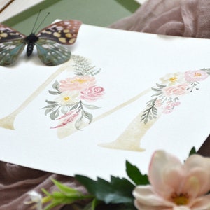 May include: Watercolor painting of the letter M decorated with pink and white flowers and green leaves.