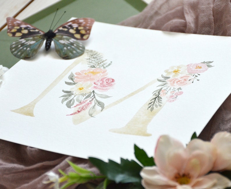 May include: Watercolor painting of the letter M decorated with pink and white flowers and green leaves.
