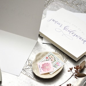 May include: A white card with the text "Will you be my junior bridesmaid?" written in purple calligraphy. The card is on a silver tray with a small plate of vintage stamps.
