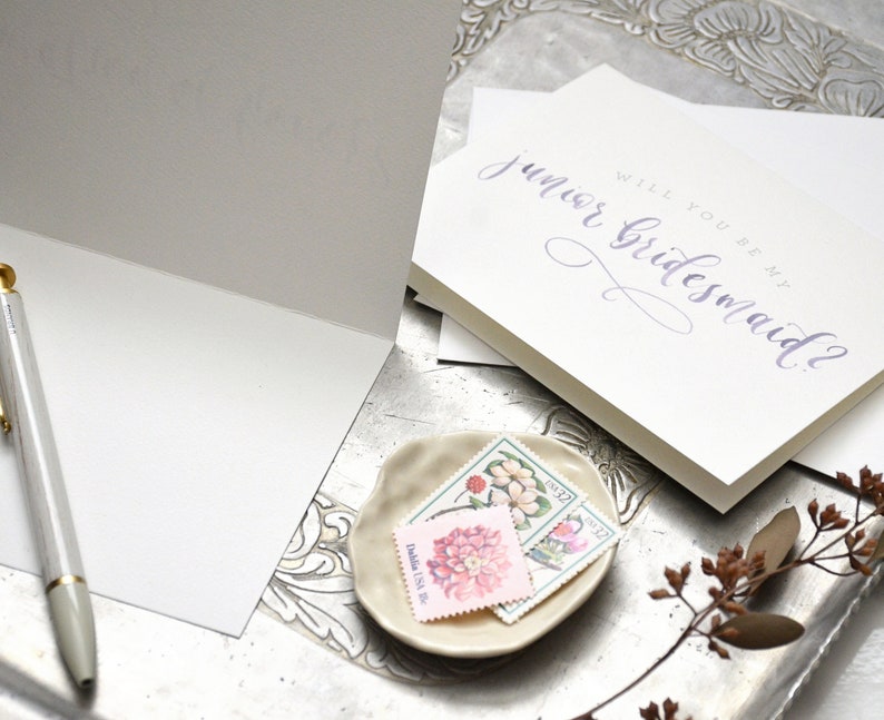 May include: A white card with the text "Will you be my junior bridesmaid?" written in purple calligraphy. The card is on a silver tray with a small plate of vintage stamps.